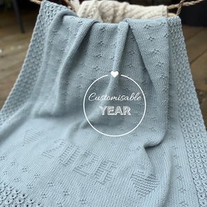 Puede incluir: Una manta de punto azul claro con un borde decorativo y la palabra "YEAR" en un círculo. La manta tiene un patrón de corazones y diamantes. La manta está doblada sobre una cesta.