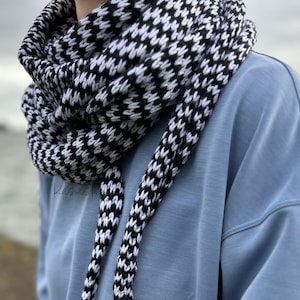 Puede incluir: Una bufanda infinita de patrón blanco y negro con un diseño de pata de gallo. La bufanda se lleva alrededor del cuello de una persona que lleva una camisa azul claro.