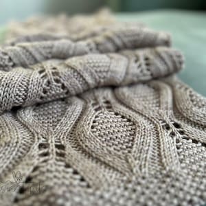 May include: A beige knitted scarf with a cable knit pattern and a delicate lace design. The scarf is folded in half and the text "Kasias Knits" is visible in the bottom left corner.