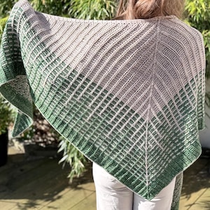 May include: A knitted shawl in shades of green and gray, draped over the shoulders. The shawl features a geometric pattern with a gradient effect, transitioning from light gray to a deep green. The texture appears soft and cozy.