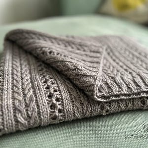 May include: A folded, gray knitted blanket with intricate cable and lace patterns. The blanket is made of soft yarn and has a textured appearance. The blanket is folded on a green surface. The blanket is a cozy home decor item.