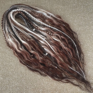 Op de afbeelding: Een verzameling bruine, witte en grijze dreadlocks met een golvende textuur. De dreadlocks zijn verweven met vlechten en decoratieve kralen. Het kleurenpalet omvat tinten bruin, wit en grijs, waardoor een natuurlijk en gestructureerd uiterlijk ontstaat.