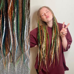 May include: A collection of colorful dreadlocks in shades of green, brown, white, and gold. The dreadlocks are shown in a close-up view, with some hanging down. The dreadlocks are a mix of colors and textures.