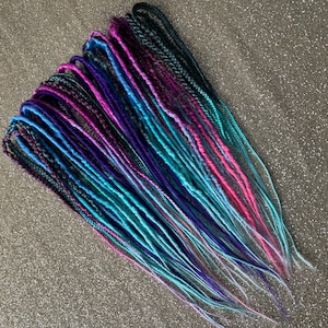 May include: A bundle of colorful dreadlocks in shades of teal, blue, purple, and pink. The synthetic hair extensions are arranged in a fan shape, with the tips of the dreads fading to a lighter color. The dreadlocks are designed for braiding into the hair.