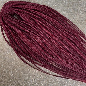 Può includere: Un fascio di capelli sintetici intrecciati color bordeaux. I capelli sono strettamente intrecciati, con ogni treccia uniforme per dimensioni e forma. I fili sono disposti in un motivo fluido e stratificato, creando un aspetto pieno e strutturato. I capelli hanno una leggera lucentezza.
