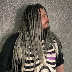 Könnte beinhalten: Eine Person mit langen, grauen Dreadlocks und Bart, trägt einen schwarzen Sweatshirt mit einem weißen Skelett-Design. Der Hintergrund ist eine glitzernde, silberne Oberfläche. Das Skelett-Design enthält ein lila Herz.