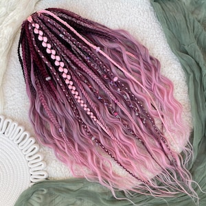 Peut inclure: Ensemble de fausses dreadlocks synthétiques roses et violettes. Les dreads sont coiffées avec des tresses, des sections ondulées et des perles décoratives. Les cheveux sont disposés sur une surface blanche avec un tissu vert en arrière-plan.