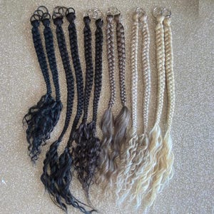 May include: A selection of braided hair extensions in shades of black, brown, grey, and blonde. Each extension has a metal ring at the top for attachment and wavy ends. The extensions are presented against a glittery background.