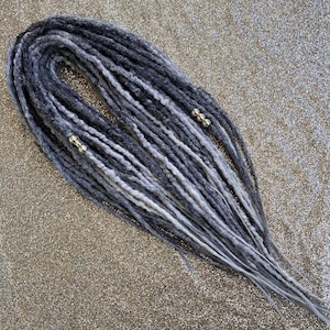 May include: A bundle of gray and white synthetic dreadlocks. The dreads are a mix of dark gray, light gray, and white, with a few silver-colored beads attached. The dreadlocks are arranged in a loose, flowing pattern.