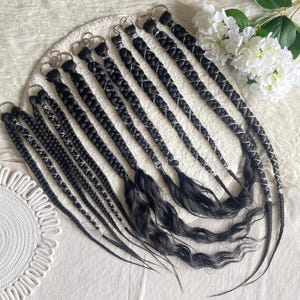May include: A collection of black synthetic hair extensions featuring intricate braids wrapped with silver metallic thread. Each extension is attached to a circular metal ring at the top, with the braids transitioning into loose, wavy hair ends.