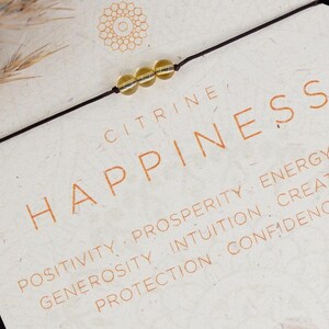 Könnte beinhalten: Eine weiße Karte mit dem Wort "Happiness" in orangefarbener Schrift. Die Karte enthält auch eine Liste positiver Affirmationen, die mit dem Wort "Happiness" verbunden sind. Die Karte zeigt ein kleines Stück Citrin-Kristall und die Wörter "Citrine" und "Air" in orangefarbener Schrift. Die Karte hat einen dekorativen Rand.