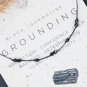 May include: Black tourmaline grounding necklace with black beads on a black cord. The card reads "Black Tourmaline Grounding" and lists the benefits of the stone, including protection, confidence, calm, balance, determination, clarity, and positivity. The card also states "Root Chakra" and "Earth".