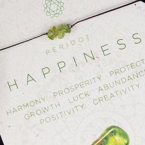 Könnte beinhalten: Eine weiße Karte mit grünen Peridot-Kristallen und dem Text "PERIDOT HAPPINESS" und "HARMONY PROSPERITY PROTECTION GROWTH LUCK ABUNDANCE POSITIVITY CREATIVITY". Die Karte enthält auch den Text "ELEMENT EARTH HEART CHAKRA SOLAR PLEXUS CHAKRA".