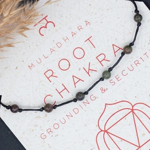 Puede incluir: Un brazalete de cuerda negra con cuentas verdes y marrones. El brazalete está etiquetado como "Muladhara Root Chakra Grounding & Security". La tarjeta también incluye un símbolo rojo y texto sobre las propiedades del chakra.
