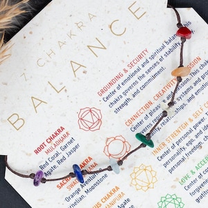 Può includere: Un braccialetto per l'equilibrio dei chakra con pietre preziose colorate è esposto su una carta con informazioni sui chakra. La carta contiene testo su radicamento, sicurezza, connessione, creatività e altri concetti di chakra. Lo sfondo è neutro.