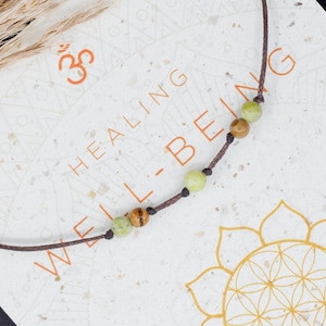 May include: A brown cord necklace with green and brown beads, on a white card with the text "HEALING WELL-BEING" and a flower symbol. The card also has the text "BALANCE HAPPINESS MANIFESTATION INNER PEACE ABUNDANCE JOY POSITIVITY ENERGY LOVE".
