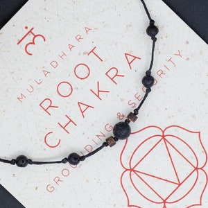 Peut inclure: Un bracelet en pierre de lave noire et cordon noir avec un symbole rouge et blanc. La carte indique "Muladhara Root Chakra Grounding & Security" et énumère les bienfaits du chakra. La carte comprend également une offre spéciale pour l'achat de plusieurs articles.