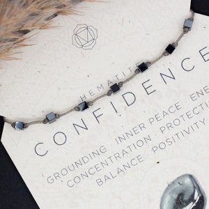 Può includere: Una collana di pietre di ematite con perline quadrate è esposta su una carta con la parola "CONFIDENCE" e il testo "GROUNDING INNER PEACE ENERGY CONCENTRATION PROTECTION BALANCE POSITIVITY". La carta presenta anche il testo "EARTH & FIRE ROOT CHAKRA".
