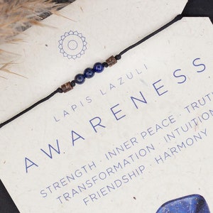 Op de afbeelding: Een blauwe lapis lazuli armband met een bruin koord. De kaart vermeldt "Lapis Lazuli Bewustzijn" en somt de eigenschappen van de steen op: "Kracht, Innerlijke Vrede, Waarheid, Transformatie, Intuïtie, Vriendschap, Harmonie". De kaart vermeldt ook "Water, Keelchakra & Derde Oog Chakra".