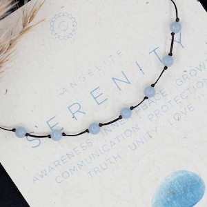 Peut inclure: Un bracelet en pierre d'angélite bleu clair avec un cordon marron. Le bracelet est sur une carte avec les mots "SERENITY" et "AWARENESS INNER PEACE GROWTH COMMUNICATION PROTECTION TRUTH UNITY LOVE" imprimés dessus. La carte a également une image de pierre bleu clair ressemblant à une aquarelle et le mot "AIR" imprimé dessus.