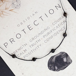 May include: Black obsidian necklace with a black cord and silver beads. The necklace is on a white card with the text "Obsidian Protection" and "Strength, Grounding, Courage, Calm, Truth, Purification, Positivity, Confidence". The card also says "Earth & Fire" and "Root Chakra, Solar Plexus Chakra".