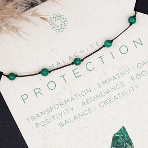 May include: A green malachite stone necklace with a brown cord. The card reads "Malachite Protection" and lists the stone's properties: transformation, empathy, calm, positivity, abundance, focus, balance, and creativity. The card also states "Earth Heart Chakra".