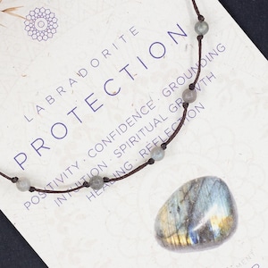 Puede incluir: Un collar de cordón marrón con cuentas de labradorita y un colgante de labradorita más grande. La tarjeta dice "Labradorita Protección" y enumera los beneficios de la piedra, que incluyen "positividad, confianza, conexión a tierra, intuición, crecimiento espiritual, sanación, reflexión". La tarjeta también dice "Aire, Chakra del tercer ojo, Chakra de la corona".