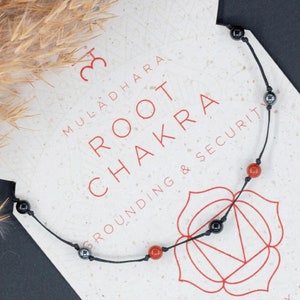 Könnte beinhalten: Ein schwarzes Kordelarmband mit roten Edelsteinperlen und schwarzen Hämatitperlen. Das Armband wird auf einer weißen Karte mit dem Text "MULADHARA ROOT CHAKRA ROUNDING & SECURITY" und einem roten und weißen Blumensymbol präsentiert. Die Karte enthält auch Text über die Vorteile des Wurzelchakras, wie z. B. Stärke, Positivität, Entschlossenheit, Schutz, Mut, Reinheit, Integrität, Vertrauen, Ruhe und Erde.