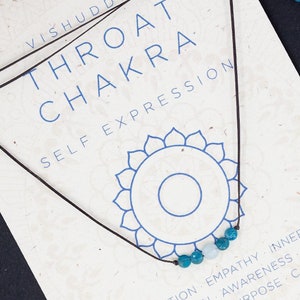 Könnte beinhalten: Eine schwarze Schnurkette mit türkisfarbenen Perlen, ein Symbol des Halschakras, und dem Text "VISHUDDHA THROAT CHAKRA SELF EXPRESSION" auf einer weißen Karte. Die Karte enthält auch eine Beschreibung der Bedeutung des Chakras und der zugehörigen Körperteile.