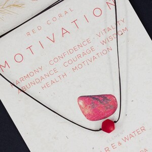 May include: A red coral necklace with a black cord, displayed on a card with the word "MOTIVATION" and related words. The card also features the text "FIRE & WATER" and "ROOT CHAKRA". Scattered red beads are in the background.