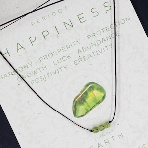May include: A green peridot stone necklace on a black cord with the words "PERIDOT HAPPINESS" and "EARTH HEART CHAKRA & SOLAR PLEXUS CHAKRA" printed on a white card. The card also includes a list of words associated with the stone, such as "harmony, prosperity, protection, growth, luck, abundance, positivity, creativity".