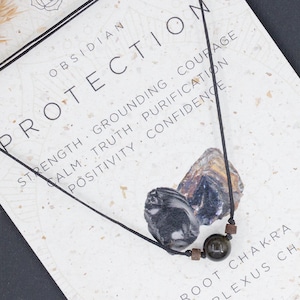 Può includere: Una collana di pietra di ossidiana nera con un cordoncino nero. La carta recita "Obsidian Protection" e elenca i benefici della pietra, tra cui forza, radicamento, coraggio, calma, verità, purificazione, positività e fiducia. La carta elenca anche il chakra radice e il chakra del plesso solare. La carta è su una superficie grigio scuro.