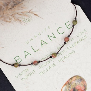 May include: A brown cord necklace with green and red beads, a pink and green stone, and a card that reads "UNAKITE BALANCE VISION GROUNDING BIRTH LOVE INSIGHT GROWTH PERSEVERANCE RELEASE HEALING FIRE & WATER HEART CHAKRA THIRD EYE CHAKRA".