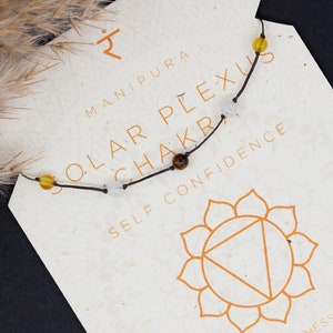 May include: A black cord bracelet with yellow, white, and brown beads. The bracelet is labelled "Solar Plexus Chakra" and "Self Confidence". The card also includes the Sanskrit symbol for the Manipura chakra and a graphic of the chakra.