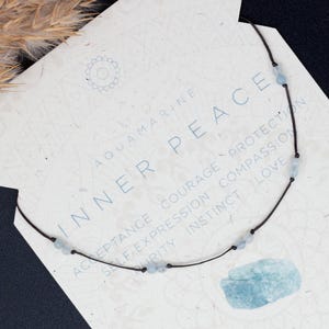 Peut inclure: Un délicat collier en aigue-marine avec un cordon noir repose sur une carte blanche avec les mots "Inner Peace" et "Aquamarine" imprimés. La carte contient également le texte "Cadeau gratuit pour chaque achat" et "Économisez plus sur les ensembles assortis!"
