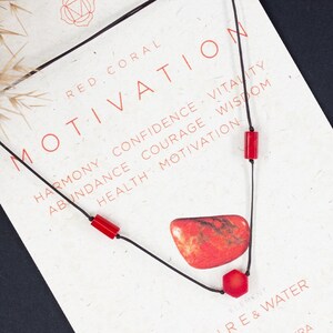 May include: Red coral necklace with a red stone pendant and black cord. The card reads "Red Coral Motivation" and lists the benefits of red coral, including harmony, confidence, vitality, abundance, courage, wisdom, health, and motivation. The card also says "Fire & Water" and "Root Chakra".