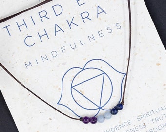 May include: A Third Eye Chakra necklace with a black cord and purple and blue beads, displayed on a card with text about mindfulness and spirituality. The card has a blue chakra symbol and the text "AJNA".