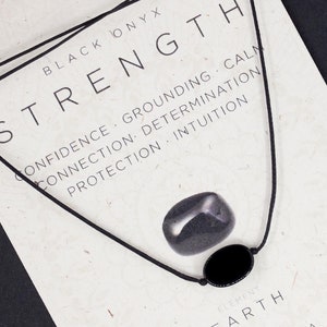 Op de afbeelding: Een zwarte onyx ketting met een zwarte ovale hanger en een zwarte steen, weergegeven op een kaart met het woord "STRENGTH" en gerelateerde zoekwoorden. De kaart bevat ook tekst over de wortelchakra en de aarde-elementen.