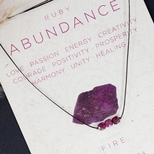 May include: A necklace featuring a red ruby stone pendant and four smaller red ruby beads. The necklace is displayed on a white card with the words "RUBY ABUNDANCE" and "FIRE" printed on it. The card also lists the benefits of ruby, including love, passion, energy, creativity, courage, positivity, prosperity, harmony, unity, and healing. The card also states that the ruby is associated with the root and heart chakras.