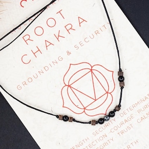 May include: A black cord necklace with black and brown beads is displayed on a white card. The card features the words "ROOT CHAKRA" in red, along with a geometric design and text about grounding and security. The background is dark.