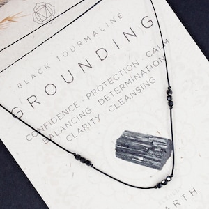Op de afbeelding: Zwarte toermalijn aarding ketting met zwarte kralen en een zwarte stenen hanger. De kaart leest "Zwarte toermalijn Aarding Zelfvertrouwen, Bescherming, Rust, Balanceren, Vastberadenheid, Duidelijkheid, Reiniging, Aarde, Wortelchakra".