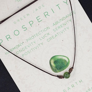 Op de afbeelding: Een groene jade ketting met een donkerbruin koord, gepresenteerd op een kaart met de woorden "Green Jade Prosperity". De kaart vermeldt kwaliteiten als harmonie, bescherming en creativiteit. Een groene jade steen en twee kralen zitten op het koord.