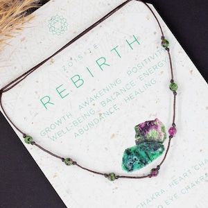 Puede incluir: Un collar de piedra verde y rosa con cordón marrón y pequeñas cuentas verdes. El collar está sobre una tarjeta blanca con el texto "ZOISITE REBIRTH" y una lista de palabras que incluyen "crecimiento", "despertar", "positividad", "bienestar", "equilibrio", "energía", "abundancia", "sanación", "chakra raíz", "chakra del corazón", "chakra del tercer ojo".