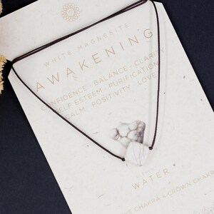 May include: A white magnesite necklace with a heart-shaped pendant, displayed on a card with the word "AWAKENING" and related keywords. The card is on a dark surface, with dried flowers in the upper left corner.