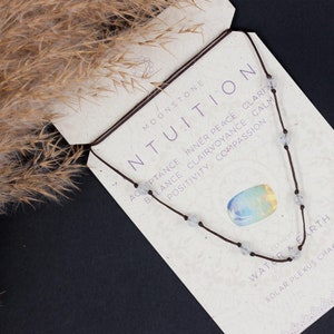 Puede incluir: Un collar de cordón marrón con cuentas de piedra lunar blancas. El collar está sobre una tarjeta con las palabras "Moonstone Intuition" y "Acceptance Inner Peace Clairvoyance Calm Balance Positivity Compassion Water & Earth Solar Plexus Chakra".