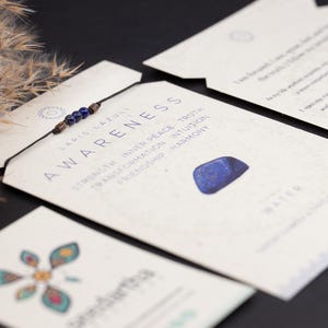 May include: A blue lapis lazuli stone bracelet with a brown cord and blue beads. The bracelet is on a card with the words "AWARENESS" and "LAPIS LAZULI" printed on it. The card also has a list of keywords: "STRENGTH, INNER PEACE, TRUTH, TRANSFORMATION, INTUITION, FRIENDSHIP, HARMONY".