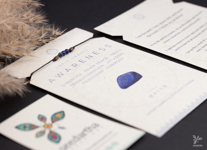 May include: A blue lapis lazuli stone bracelet with a brown cord and blue beads. The bracelet is on a card with the words "AWARENESS" and "LAPIS LAZULI" printed on it. The card also has a list of keywords: "STRENGTH, INNER PEACE, TRUTH, TRANSFORMATION, INTUITION, FRIENDSHIP, HARMONY".