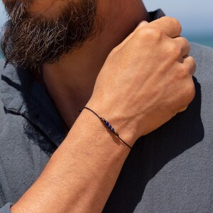 May include: A simple bracelet with a black cord and small blue beads. The bracelet is worn on a person's wrist.