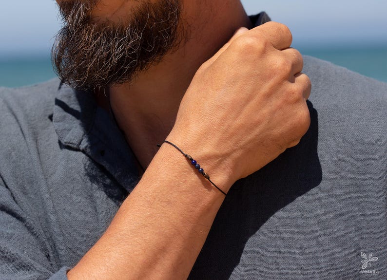 May include: A simple bracelet with a black cord and small blue beads. The bracelet is worn on a person's wrist.