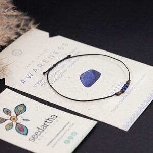 May include: A black string bracelet with blue lapis lazuli beads and a small lapis lazuli stone charm. The bracelet is on a card with the text "AWARENESS" and "WATER" on it.
